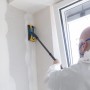 Mann in Schutzkleidung schleift Trockenbauwand mit ausziehbarem wolfcraft Teleskopstiel für Rundschleifer.