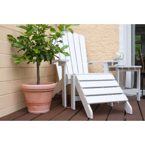 Weißer Adirondack Gartenstuhl mit Fußhocker auf Holzterrasse. Klassischer Gartensessel in Holzoptik.