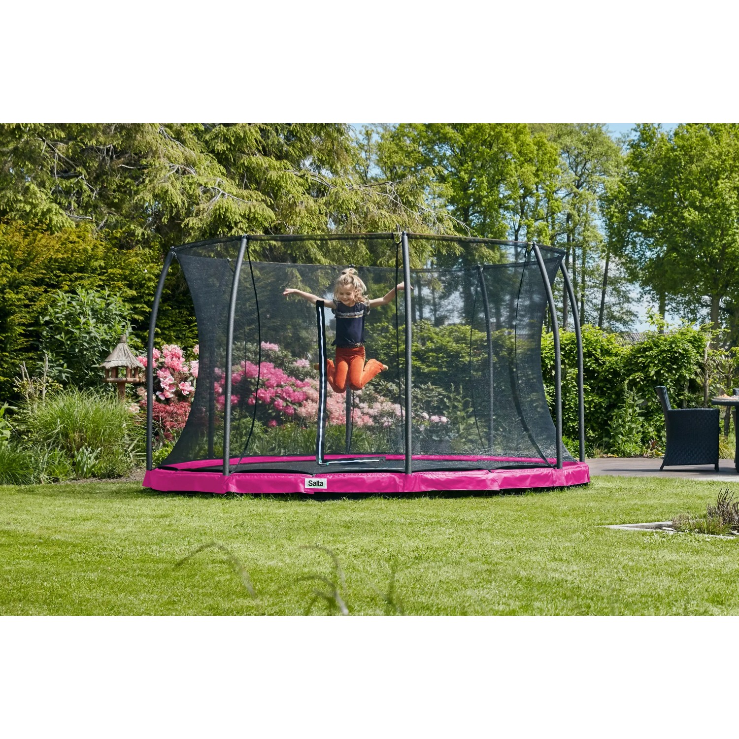 Pinkes Salta Bodentrampolin Comfort Edition (ø 251 cm) mit Sicherheitsnetz und springendem Kind im Garten.