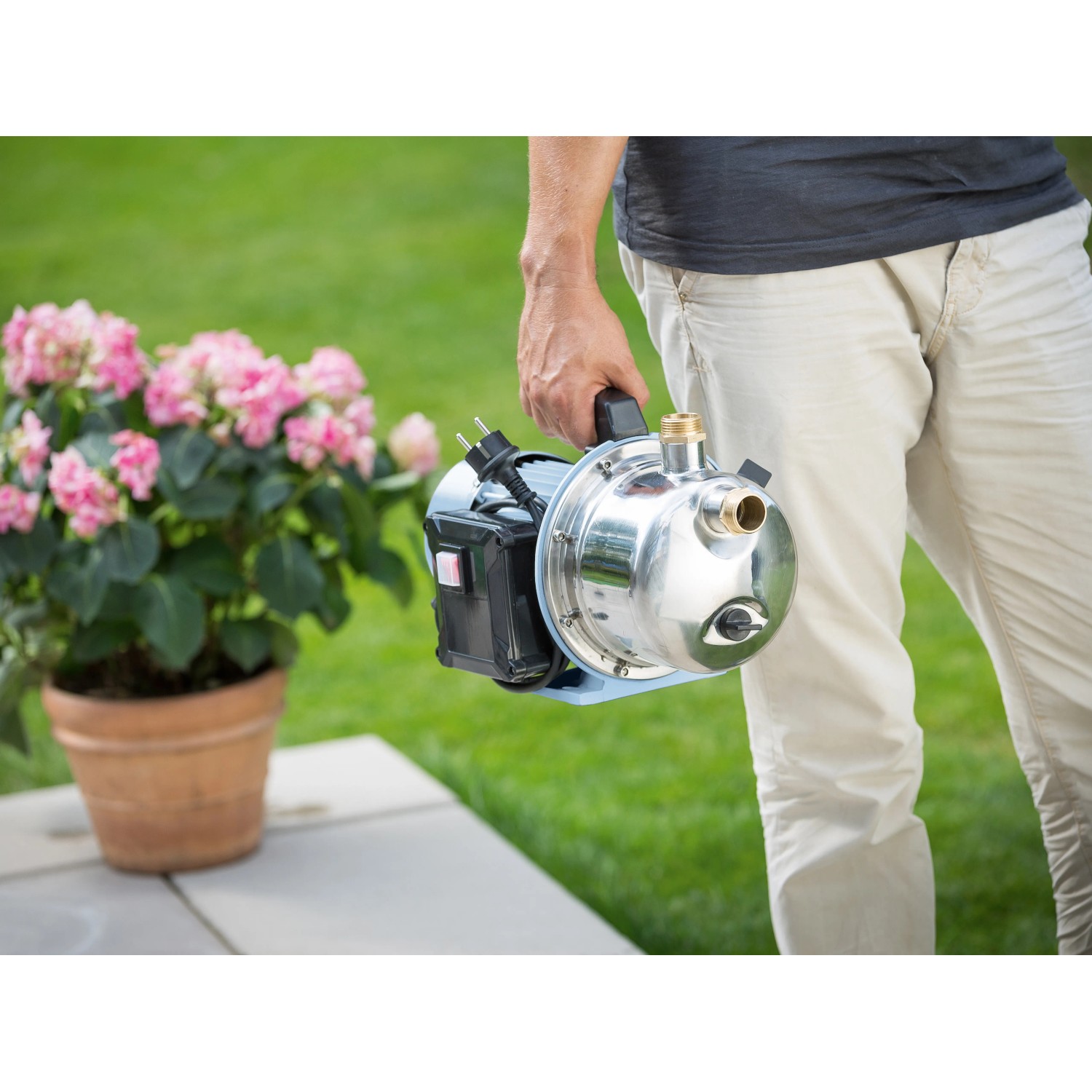 Mann trägt Gartenpumpe GP 3800E (900 W) mit Saugschlauch vor Garten. Edelstahlgehäuse.