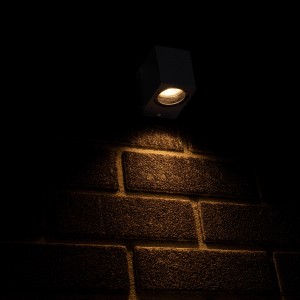 Anthrazitfarbene, eckige LED Außenwandleuchte mit Lichtkegel an einer Backsteinwand.