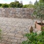 Verlegte Mehrformat-Pflastersteine Encanto Beton Antik Jura-Nuanciert auf einer Terrasse mit Steinmauer im Hintergrund.