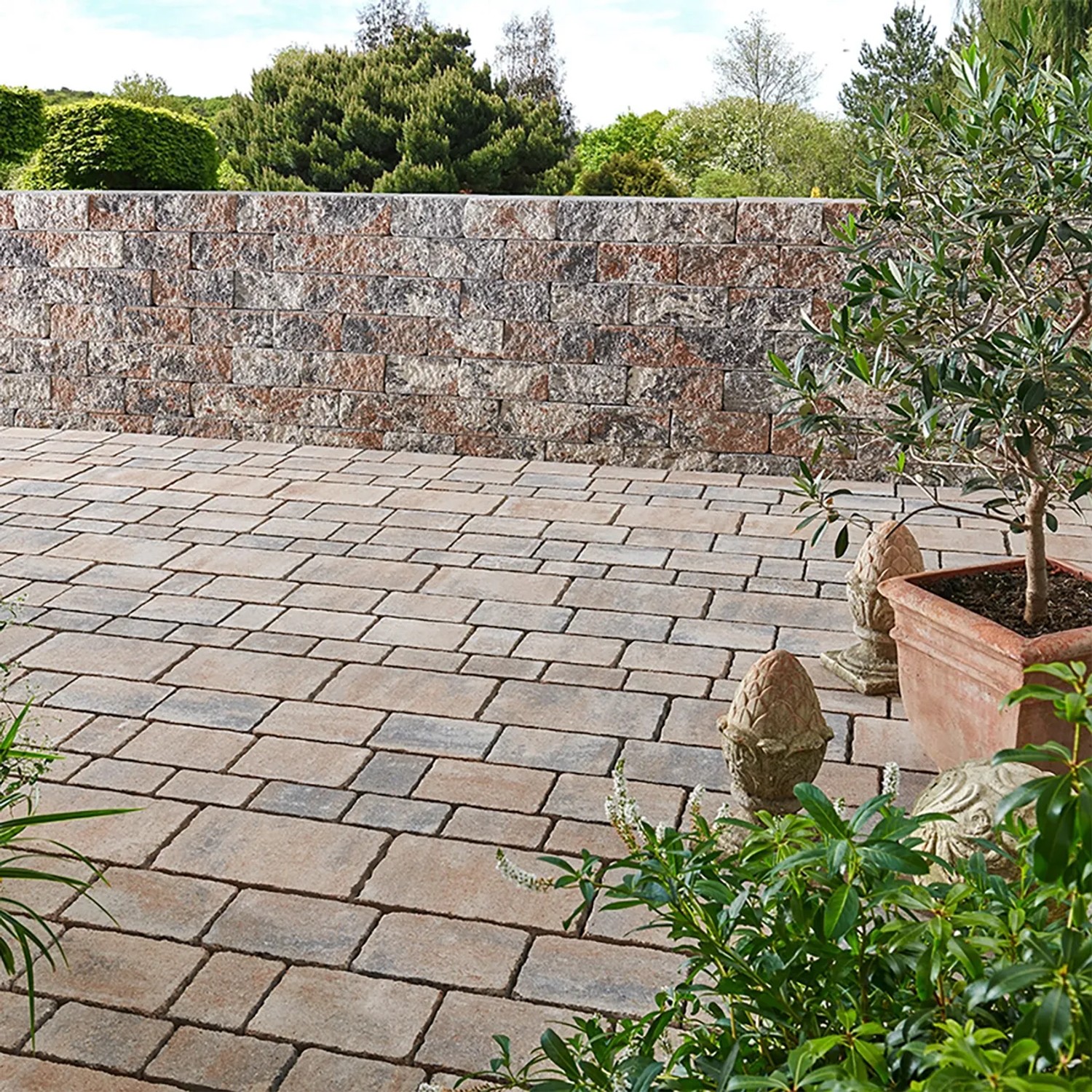 Verlegte Mehrformat-Pflastersteine Encanto Beton Antik Jura-Nuanciert auf einer Terrasse mit Steinmauer im Hintergrund.
