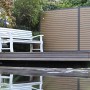GartenLand WPC Zaun Stockholm, karamellbraun, mit Bank auf Holzsteg am Wasser.