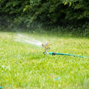 Impulsregner aus Metall und Kunststoff bewässert grünen Rasen. Der Kreisregner sprüht Wasser, angeschlossen an einen Gartenschlauch.