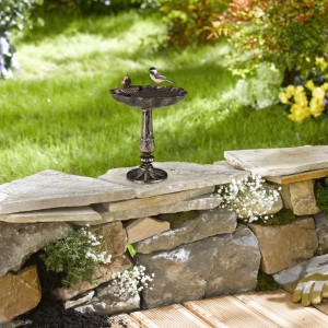 Bronzefarbene Gusseisen Vogeltränke im Muschel-Design mit Spatz-Figur und echtem Vogel auf Steinmauer im Garten.