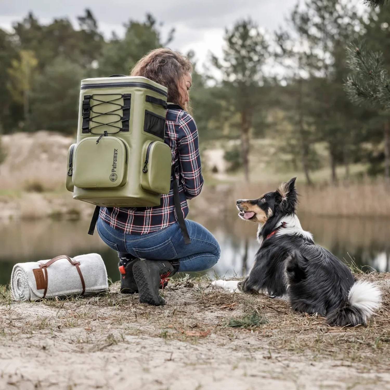 Frau mit olivgrünem Petromax Kühlrucksack 7l, Hund und Picknickdecke am See.