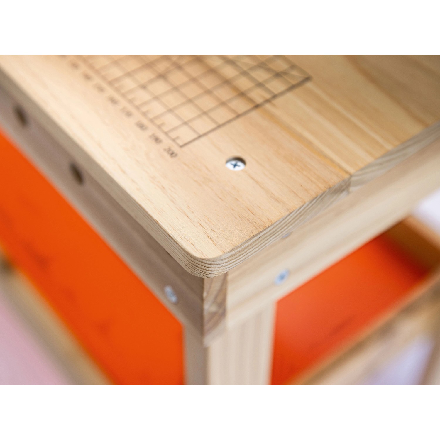 Detailansicht der OBI Kinderwerkbank aus Holz: helle Holzplatte mit aufgedrucktem Raster, abgerundete Kanten, orangefarbene Ablage.