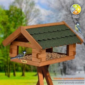 Braunes Blockhaus-Vogelhaus aus Kiefernholz mit grünem Bitumendach auf Ständer, gefüllt mit Vogelfutter. Zwei Meisen.