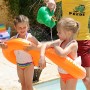 Zwei Kinder mit orangen Freds Swim Academy Schwimmreifen im Pool, ein Erwachsener gießt Wasser.