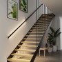 Paulmann MaxLED Stair LED-Streifen beleuchten stilvoll eine moderne Holztreppe mit schwarzem Geländer. Warmweißes Licht unter den Stufen und am Handlauf.