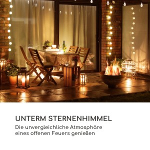 Blumfeldt Savage Feuerschale mit Grill im Garten bei Nacht. Feuer brennt, Tisch und Stühle stehen bereit.