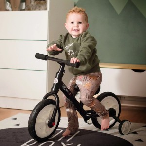 Kind auf schwarzem HOMCOM Kinderfahrrad 2-in-1 mit Stützrädern im Zimmer.