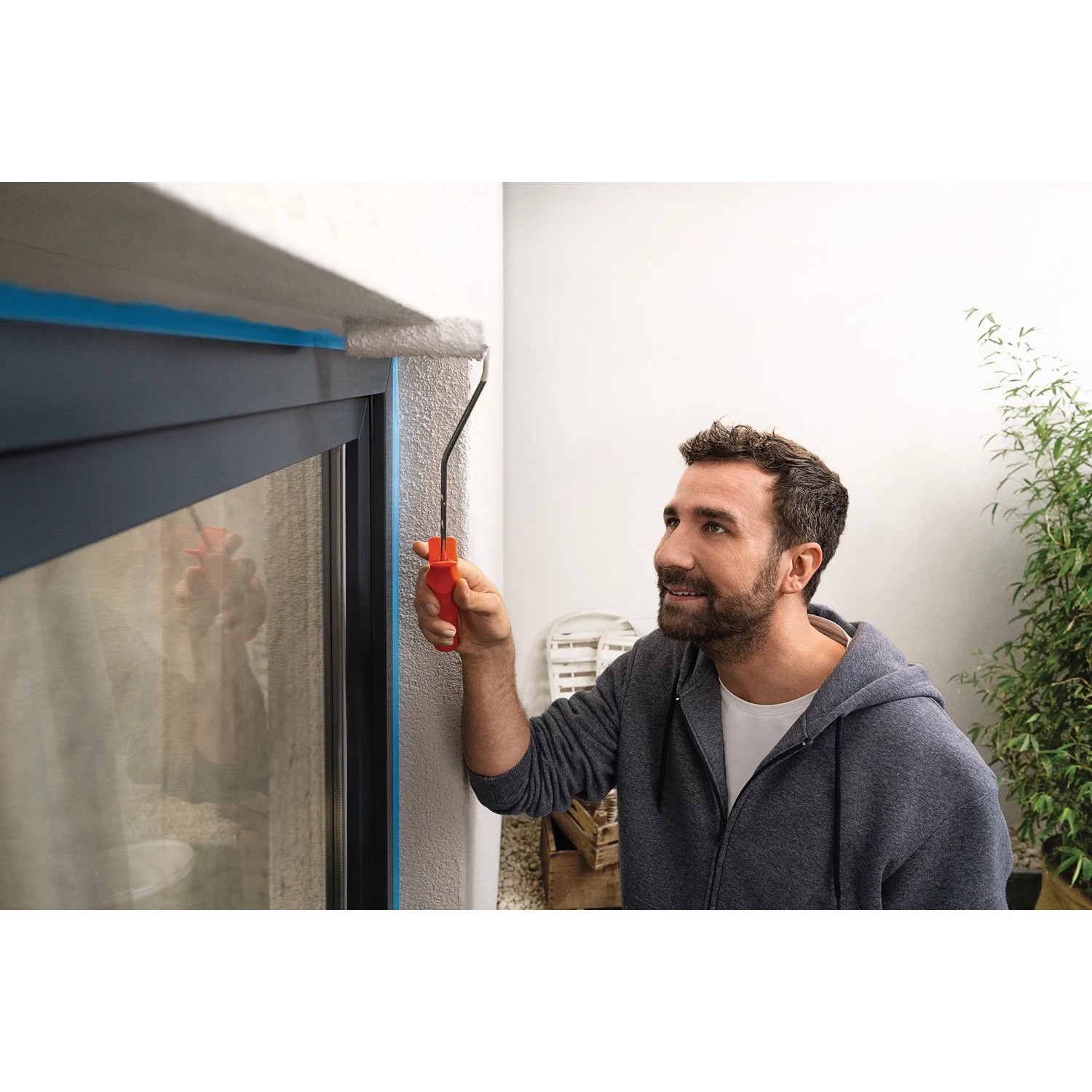 Mann streicht Fassade mit Farbroller, Fenster abgeklebt mit Tesa Malerband Professional Outdoor.