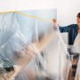 Frau bringt Tesa Abdeckfolie mit Klebeband Easy Cover Perfect+ M an Wand an.