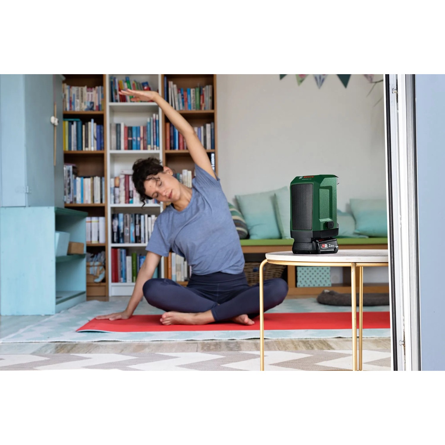 Grüner Bosch Akku-Bluetooth-Box UniversalSpeaker 18V-1 auf Tisch, Person macht Yoga. Kabelloser Lautsprecher.