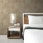 Beige Vliestapete mit barockem Ornamentmuster an einer Schlafzimmerwand, neben einem Bett und Nachttisch.