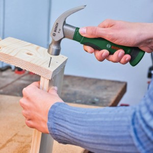 Person hämmert mit grün-silbernem Bosch DIY Klauenhammer 8oz einen Nagel in Holz. Ergonomischer Softgrip-Griff für Heimwerker.