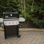 Schwarzer Weber Gasgrill E-410 Spirit mit 4 Brennern und Seitentischen vor Gartenkulisse.