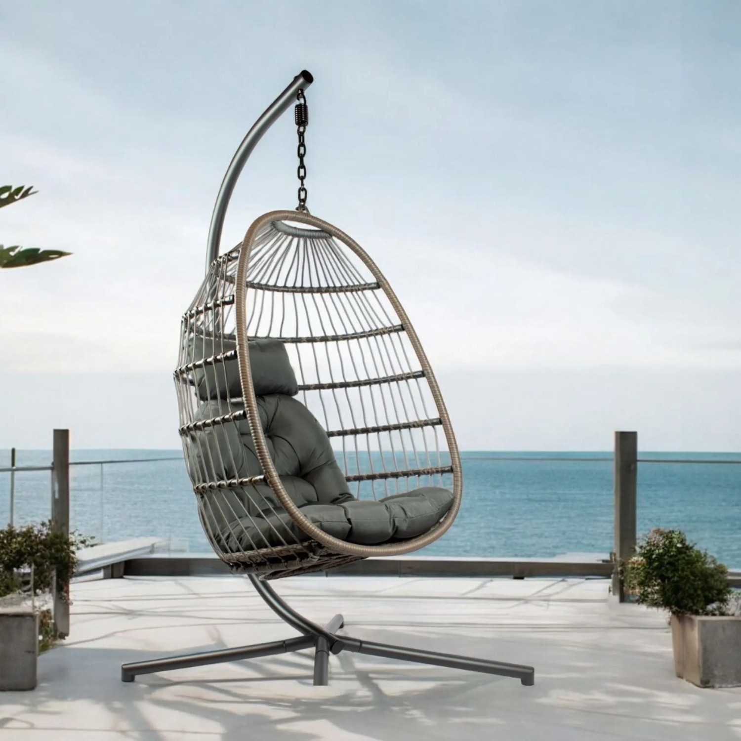 Grauer osoltus Hängesessel Pura XL mit geflochtenem Korb, Stahlgestell und Kissen auf Terrasse am Meer.