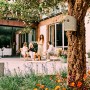 Elho Vogelhaus Cosy, beige-weiß, an Baum befestigt. Gartenszene mit Familie im Hintergrund.