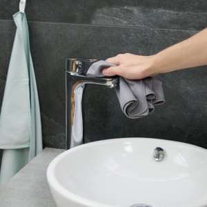 Hand reinigt Chrom-Wasserhahn mit grauem Mikrofasertuch über weißem Waschbecken. Saugstarke Putztücher für Bad.