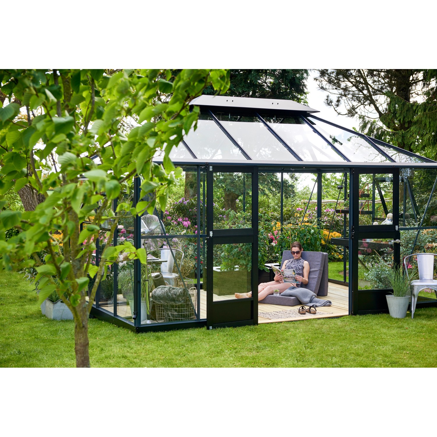 Juliana Gewächshaus Grand Oase 13 Anthrazit im Garten mit entspannender Person. Begehbares Gewächshaus mit viel Platz.