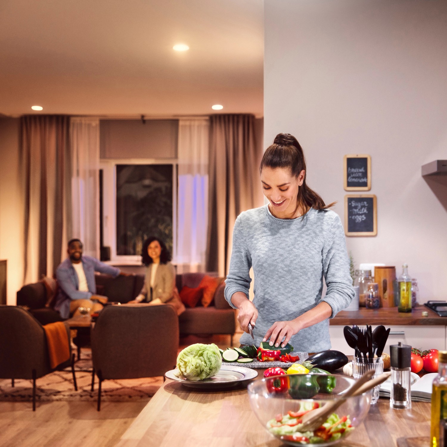 Philips Hue Starter-Set: Frau schneidet Gemüse in warmweiß beleuchteter Küche, Freunde im Hintergrund.