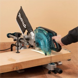 Makita Kapp- und Gehrungssäge schneidet Holzbrett. Graue Maschine mit transparentem Schutz, Staubbeutel, Hand.