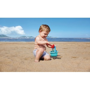 Kleinkind spielt mit Hape Babyeimer und Schaufel im Sand am Strand.