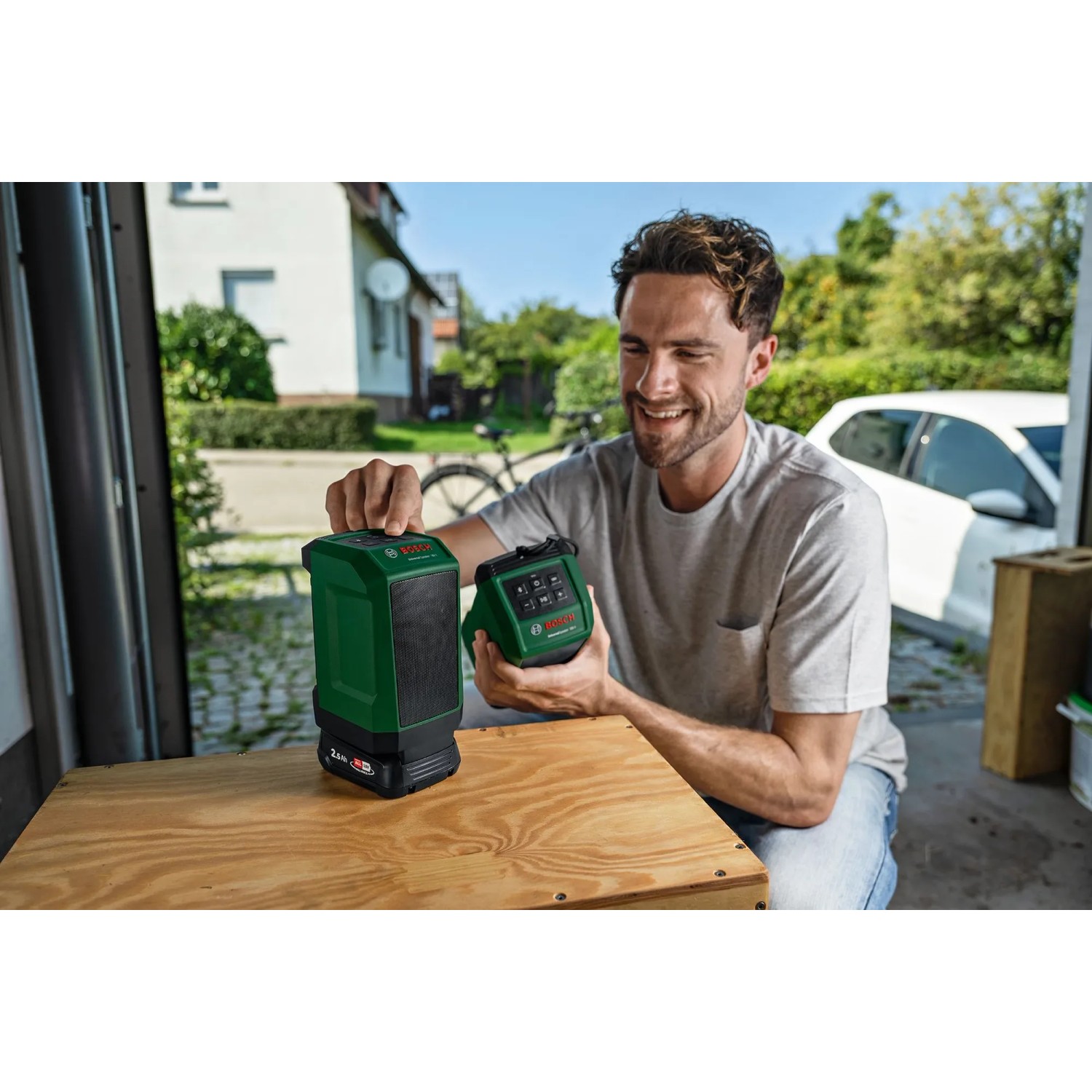 Mann hält Bosch UniversalSpeaker 18V-1 Akku-Bluetooth-Box und zweiten Lautsprecher auf Werkbank. Kompaktes Akkugerät für kabelloses Streaming.