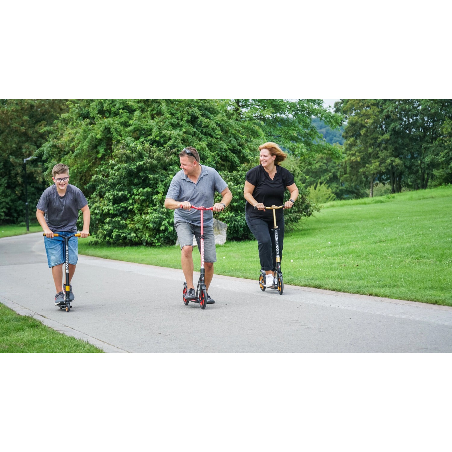 Familie fährt mit Six Degrees Aluminium Scooter (rot) auf einem Weg im Park.