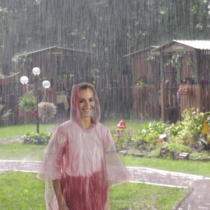 Frau im transparenten, rötlichen Polyethylen Regenponcho mit Kapuze bei starkem Regen. Praktischer Schutz für Camping & Festival.