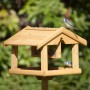 Hölzernes Vogelhaus aus Tannenholz mit Ständer, Dach und Futtersilo. Zwei Blaumeisen fressen am Futtertisch.