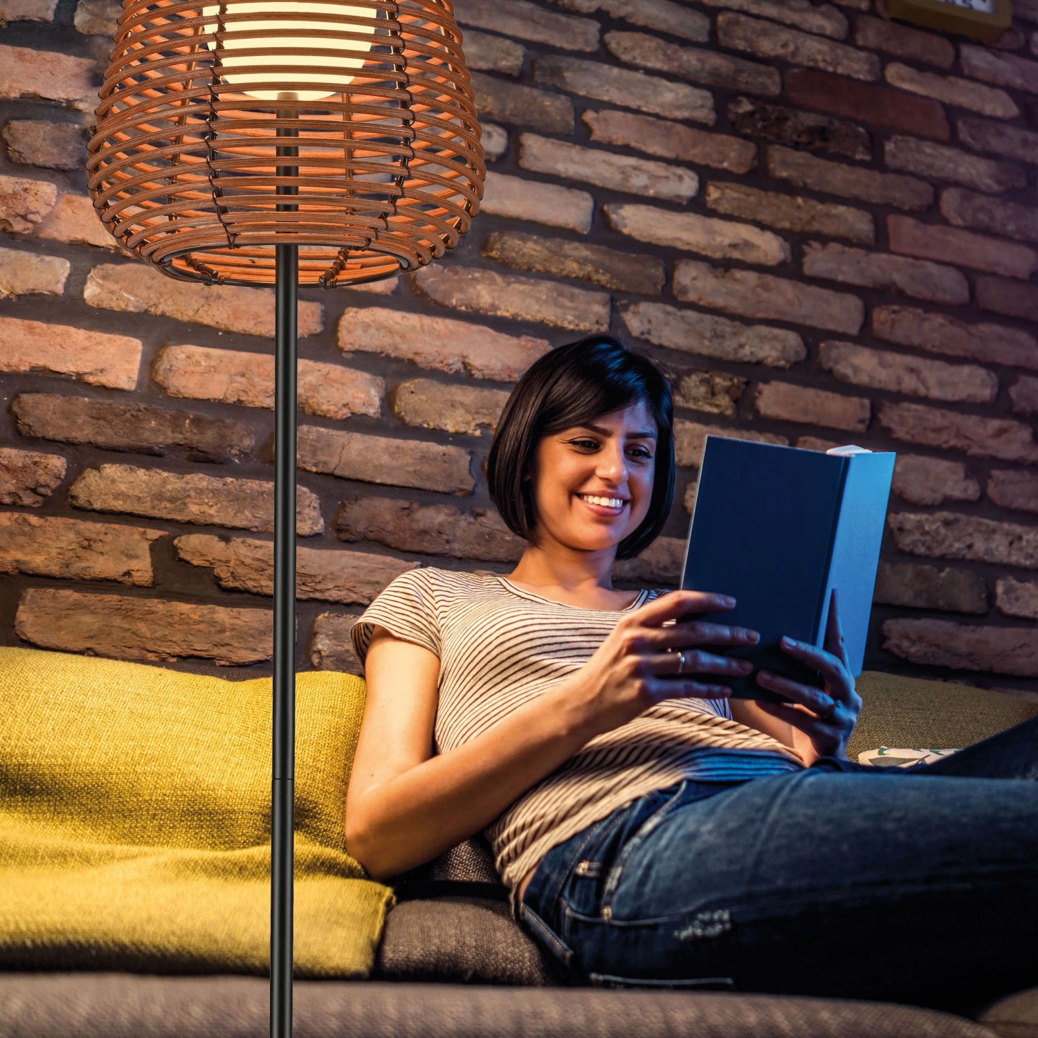 LED-Akku-Stehleuchte Askia, 120cm, schwarz, Rattan-Schirm, im Wohnraum mit lesender Person.