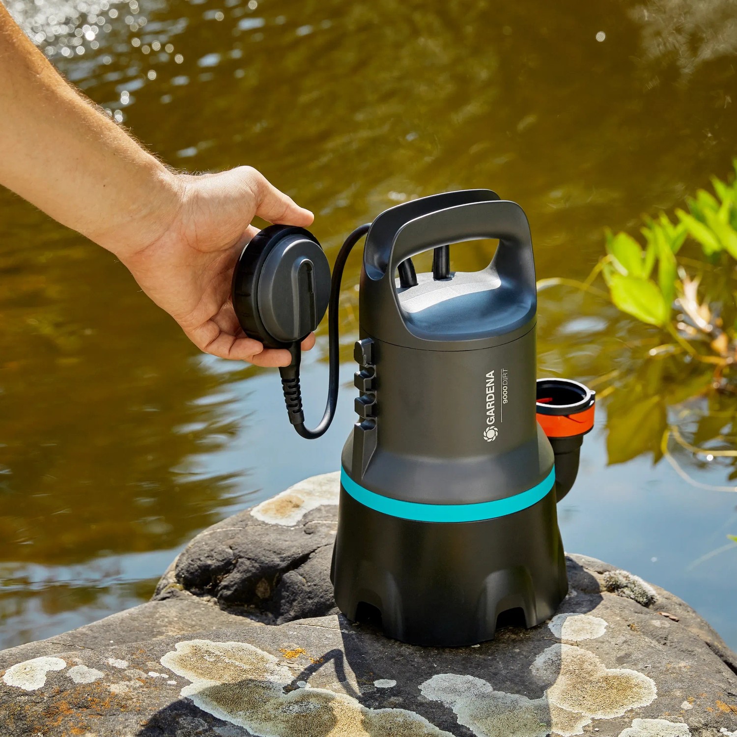 Gardena Schmutzwasser-Tauchpumpe 9000 auf Stein, mit Schwimmerschalter in der Hand vor Teich.
