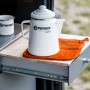 Petromax Aramid Topflappen Paar in Orange-Schwarz unter weißem Kessel und Tasse auf Schublade.