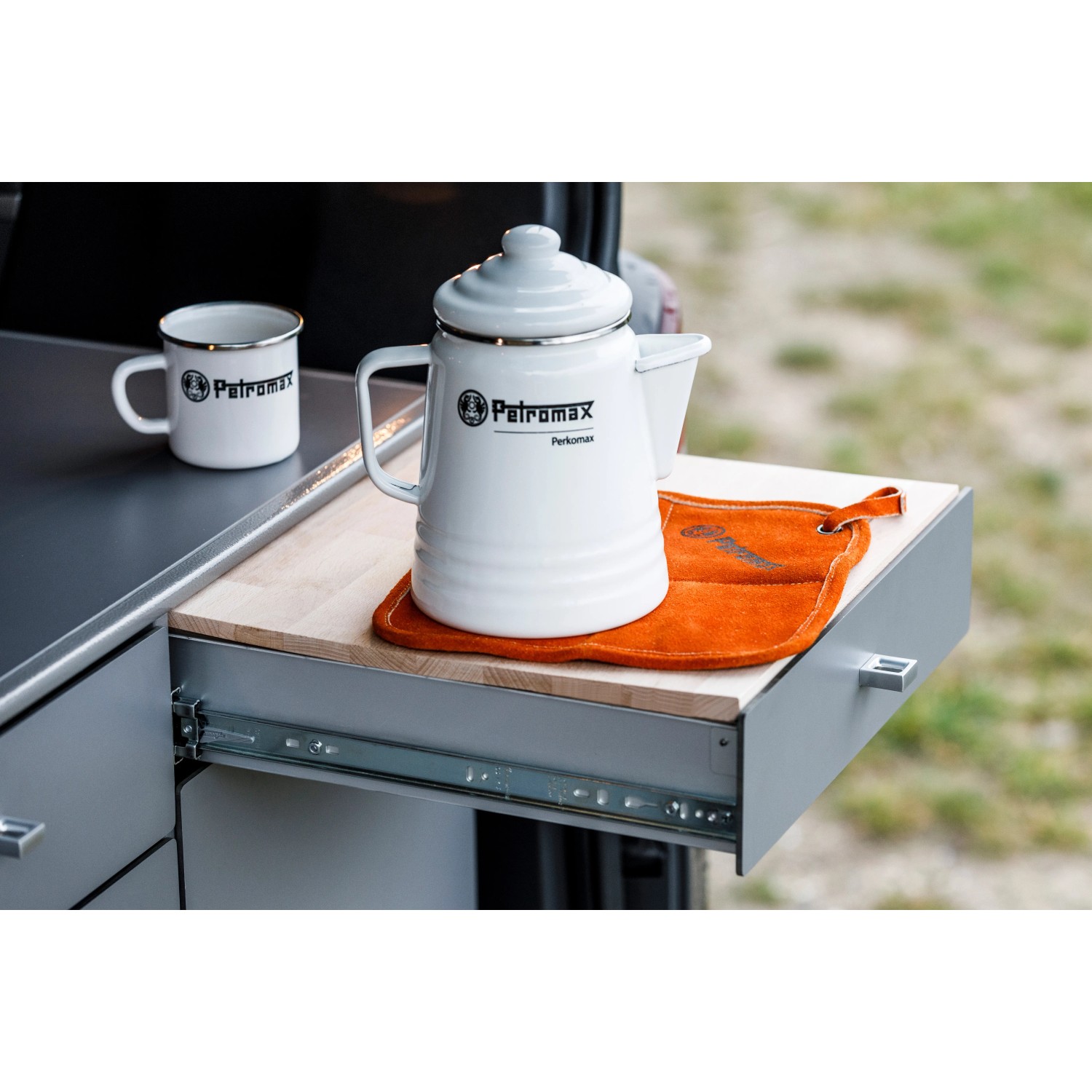 Petromax Aramid Topflappen Paar in Orange-Schwarz unter weißem Kessel und Tasse auf Schublade.