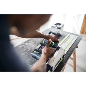 Person nutzt Festool Akku-Tauchsäge TSC55KEB-Basic 18/36 V mit Führungsschiene für präzise Holzarbeiten. Weiße Säge mit grünen Akkus.
