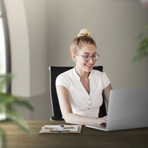 Frau sitzt lächelnd auf schwarzem Bürostuhl aus Kunstleder am Schreibtisch, arbeitet am Laptop. Ergonomischer Komfort.