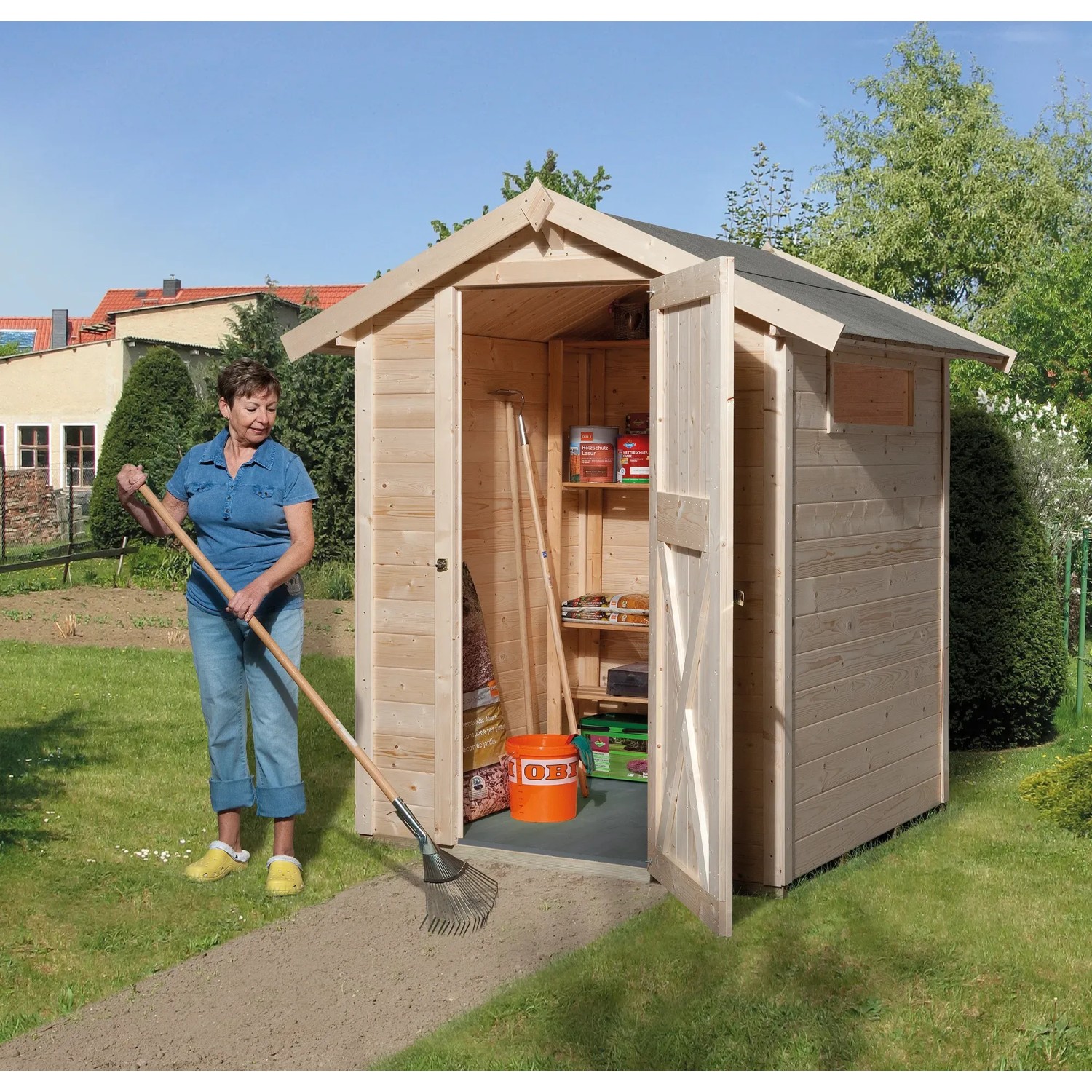 OBI Holz-Gartenhaus Kompakt mit Satteldach, 152x171 cm, naturbelassen, mit offener Tür und Frau mit Rechen.