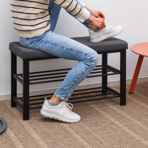 DuneDesign Schuhschrank aus Metall, grau gepolsterte Sitzbank. Person bindet Schuh. Zwei Ablagen für Schuhe.