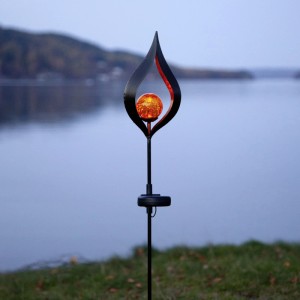 Schwarzer LED-Solarstab Flamme mit leuchtender bernsteinfarbener Glaskugel am Seeufer. Ideal für Garten-Deko.