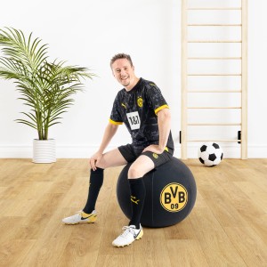 Mann sitzt auf schwarzem BVB Sitting Ball Hocker (120l) im Wohnzimmer.