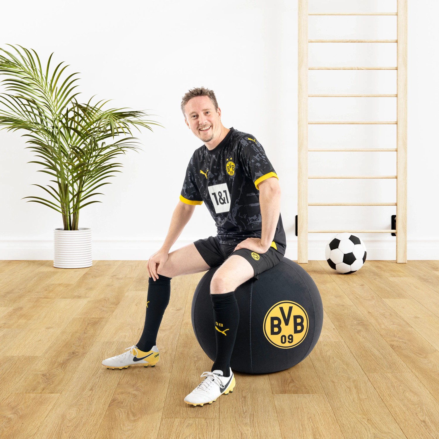 Mann sitzt auf schwarzem BVB Sitting Ball Hocker (120l) im Wohnzimmer.