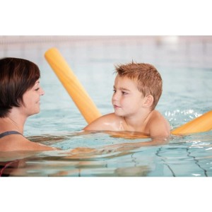 Kind mit gelber Poolnudel im Schwimmbad, eine Frau assistiert beim Schwimmenlernen.