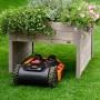 Graue Mährobotergarage mit Pflanzkasten, darunter ein Mähroboter auf Rasen.