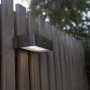 Moderne, eckige Lutec LED Solar-Wandleuchte Brick aus Edelstahl mit Bewegungsmelder an Holzzaun.