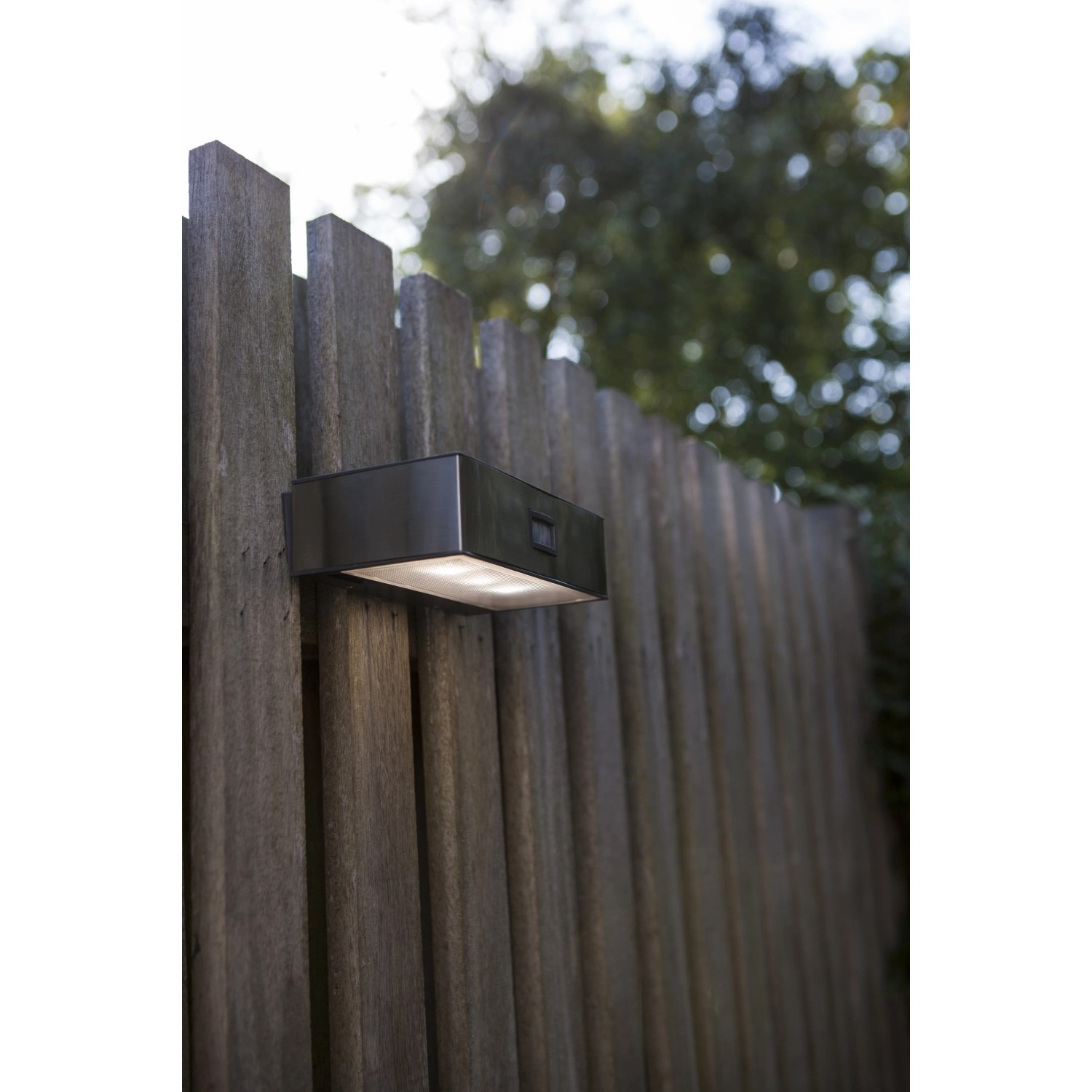 Moderne, eckige Lutec LED Solar-Wandleuchte Brick aus Edelstahl mit Bewegungsmelder an Holzzaun.