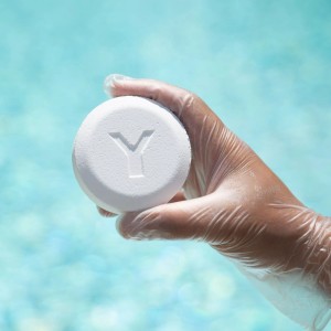 Hand mit Schutzhandschuh hält Bayrol Chlorilong Classic Chlortablette vor Poolwasser.